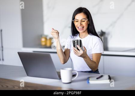 Felice donna con lo smartphone che ha ricevuto incredibili buone notizie dal messaggio sul telefono cellulare mentre lavora al computer portatile da casa, rendendo sì vincitore Foto Stock
