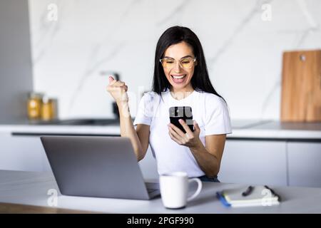 Felice donna con lo smartphone che ha ricevuto incredibili buone notizie dal messaggio sul telefono cellulare mentre lavora al computer portatile da casa, rendendo sì vincitore Foto Stock