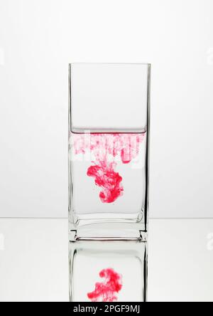 Un bicchiere d'acqua cristallino con una gocciolina rosa vibrante sospesa al centro su uno sfondo bianco Foto Stock