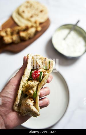 Souvlaki ona pita di pollo greco con verdure fresche e salsa tzatziki. Mano che tiene una pita con souvlaki di pollo. Sullo sfondo c'è un pollo Foto Stock