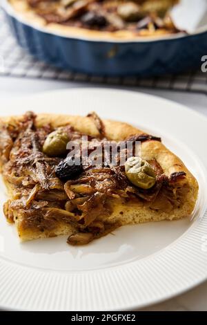 Torta di cipolle Pissaladiere con acciughe e olive nere. Il pezzo tagliato di torta si trova sul piatto, la torta rimanente nello stampo sullo sfondo Foto Stock