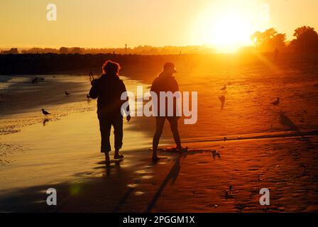 Una giovane coppia cammina sulla sabbia lungo la spiaggia in riva al tramonto Foto Stock
