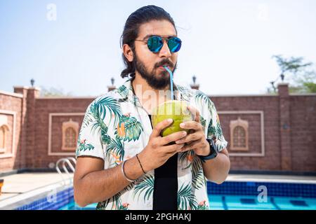 Giovane uomo indiano assetato che indossa occhiali da sole e panni casual bere acqua di cocco nel caldo giorno d'estate. Colpo di calore o onda di calore. Foto Stock