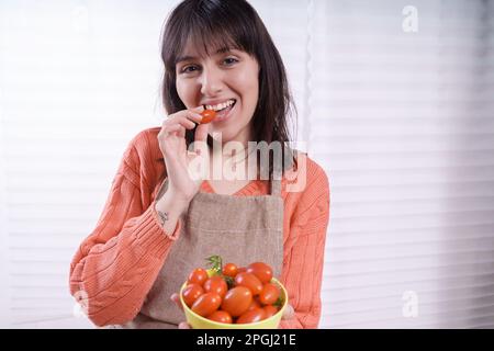 Una casalinga di brunetta caucasica con capelli lunghi in un maglione arancione e grembiule color sabbia morde un pomodoro di ciliegia, tenendo una ciotola da una finestra retroilluminata, e. Foto Stock