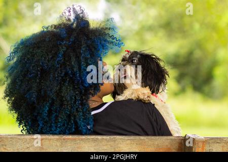 Goiania, Goias, Brasile – 20 marzo 2023: Foto di una giovane donna, dalla schiena, seduta su una panchina con un cucciolo di shitzu in grembo. Foto Stock