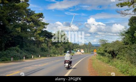 Una motocicletta guida lungo la strada AH17 nelle Highlands centrali del Vietnam vicino a buon ma Thuot accanto a una gigantesca turbina eolica. Foto Stock