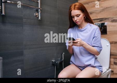 Bella giovane donna che utilizza smartphone seduto sul water in bagno di casa con interni moderni. Ritratto di donna rossa che tiene il telefono e. Foto Stock