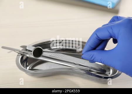 Dentista che prende lo specchio della bocca dal vassoio a forma di rene a tavola di legno, primo piano Foto Stock