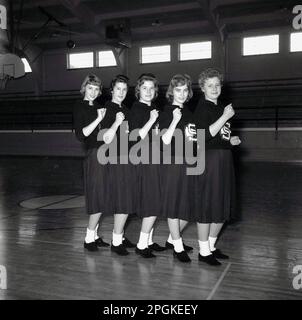 1960s, storico, all'interno di una sala sportiva, una squadra di cheerleaders nei loro abiti; top con loghi, gonne e calzini, in posa insieme per una foto, USA. Squadre di cheerleaders leader spettatori nel tifo sono una tradizione in eventi sportivi americani come il basket e il football americano. Foto Stock