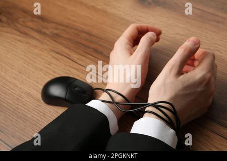 Uomo che mostra le mani legate con il cavo del mouse del computer al tavolo di legno, primo piano. Dipendenza da Internet Foto Stock