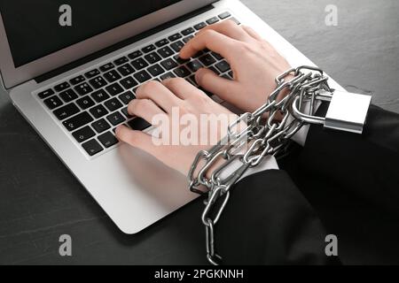 Dipendenza da Internet. Uomo che digita sul computer portatile con le mani incatenate al tavolo nero, primo piano Foto Stock