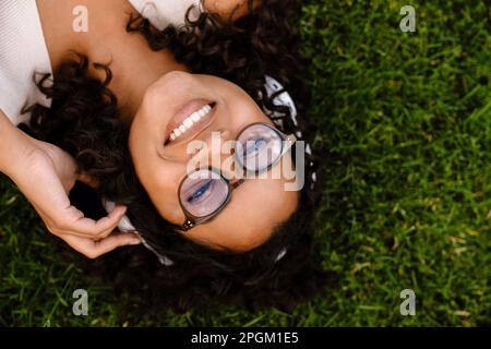 Vista dall'alto di una allegra donna africana negli occhiali che ascolta musica con le cuffie mentre riposa sull'erba nel parco Foto Stock