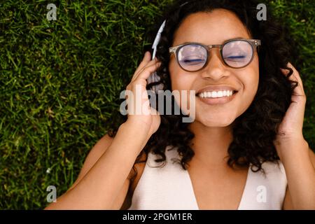 Vista dall'alto di una allegra donna africana negli occhiali che ascolta musica con le cuffie mentre riposa sull'erba nel parco Foto Stock