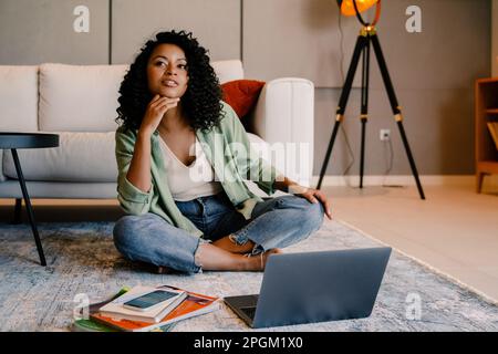 Giovane donna africana penosa che usa il notebook mentre si siede sul pavimento a casa Foto Stock