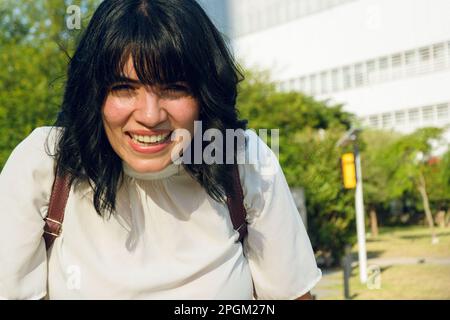molto felice giovane donna venezuelana all'aperto, ridendo guardando la macchina fotografica, indossa abiti casual e uno zaino, indossa corti capelli neri colorati, copia Foto Stock