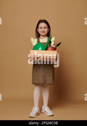 Ragazza bambino caucasica consapevole ritratto a tutta lunghezza che tiene una scatola di legno con attrezzi da giardino, isolato su sfondo beige Foto Stock