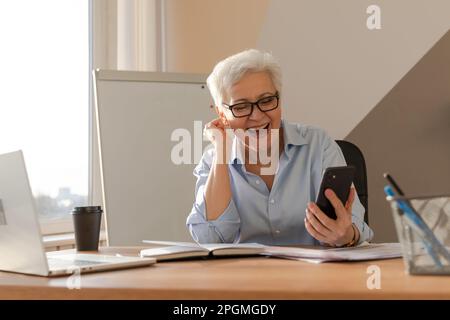 Sicura donna anziana di mezza età euforica vincitrice con smartphone. Signora più anziana matura guardando il telefono delle cellule che legge le notizie grandi ottenendo il buon risultato che vince la sensibilità in linea dell'offerta stupita. Gesto vincente Foto Stock