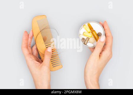 Ampolle cosmetiche con siero in mano femmina e vitamine per la crescita dei capelli, restauro capelli di bellezza. Pettine di legno con perdita di capelli su sfondo grigio. Foto Stock