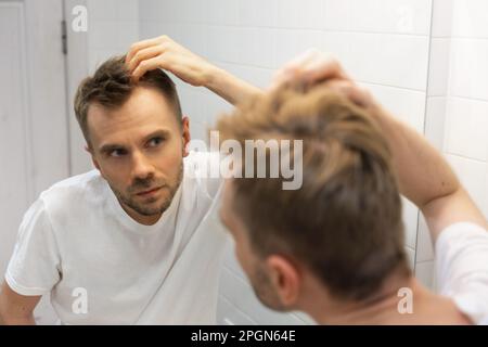 L'uomo bianco caucasico di mezza età con una barba corta guarda i capelli nello specchio del bagno e si preoccupa di calciare. Problema dei capelli dell'uomo Foto Stock