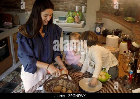 Madre che sbuccia le patate con i bambini seduti sul bancone in cucina Foto Stock
