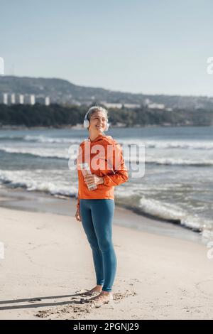 Donna matura sorridente con bottiglia d'acqua in piedi sulla riva in spiaggia Foto Stock