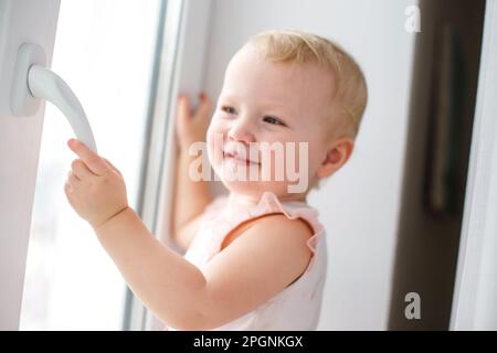 Proteggere i bambini dalla caduta dalle finestre. Una bambina attiva apre con attenzione una finestra in metallo-plastica nella casa. Foto Stock