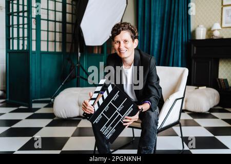 Sorridente regista seduto su sedia che tiene il clapboard al set cinematografico Foto Stock