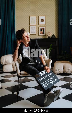 Sorridente regista seduto con clapboard su sedia al set cinematografico Foto Stock