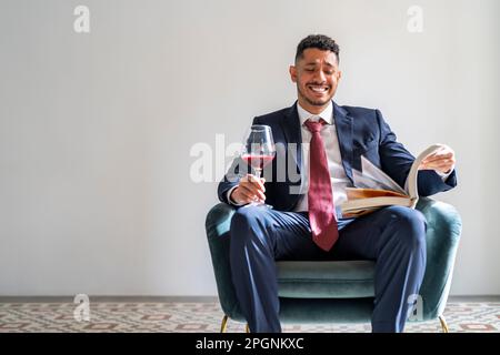 Felice uomo d'affari seduto con un libro e un bicchiere di vino rosso sulla poltrona di fronte al muro Foto Stock