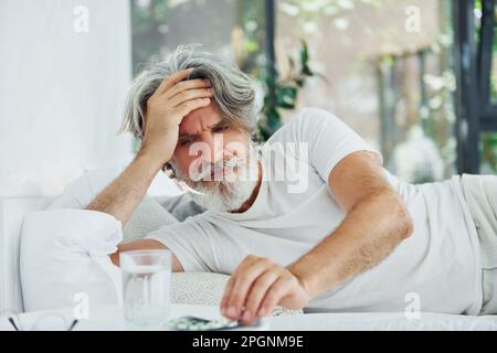 Uomo moderno ammalato di stile senior con capelli grigi e barba interna sdraiato sul letto e si sente male Foto Stock