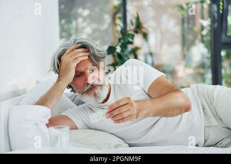 Uomo moderno ammalato di stile senior con capelli grigi e barba interna sdraiato sul letto e si sente male Foto Stock