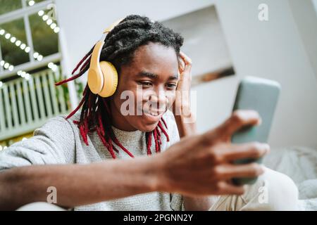 Giovane uomo sorridente che indossa le cuffie con lo smartphone a casa Foto Stock