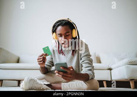 Un giovane scioccato che usa uno smartphone con carta di credito a casa Foto Stock