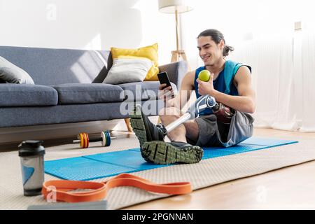 Giovane uomo sorridente con disabilità che usa uno smartphone seduto a casa Foto Stock