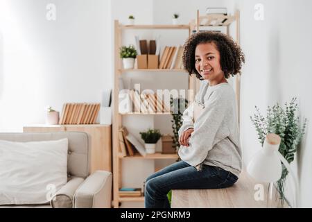 Ragazza sorridente con capelli ricci seduti sulla scrivania a casa Foto Stock