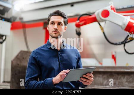 Un tecnico attento che tiene il tablet PC in piedi nella fabbrica di robot Foto Stock