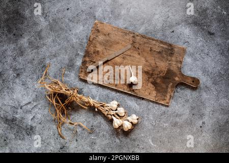 Aglio crudo, tagliere e coltello da cucina sdraiati sulla superficie grigia Foto Stock