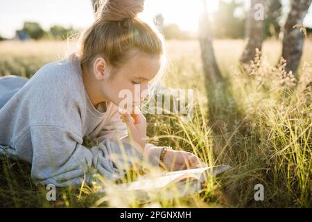 Ragazza sdraiata sull'erba e libro di lettura il giorno di sole Foto Stock