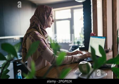 Freelance che indossa un hijab che lavora sul computer portatile in ufficio a casa Foto Stock
