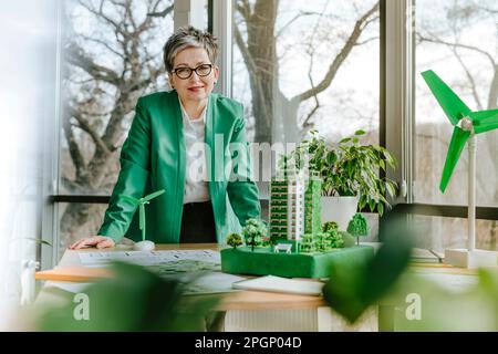 Donna d'affari sorridente in piedi con architettura biofila e modelli di turbine eoliche sulla scrivania in ufficio Foto Stock