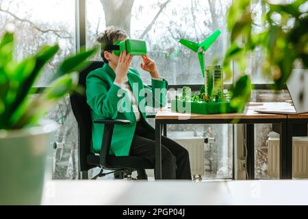 Donna d'affari che indossa occhiali VR seduti con turbina eolica e modelli architettonici biofili in ufficio Foto Stock