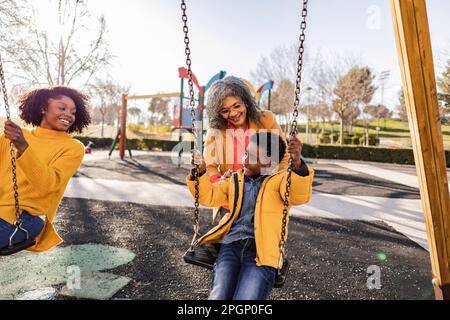 Donna sorridente che guarda la nonna che spinge il figlio sull'altalena nel parco giochi Foto Stock