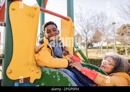 Figlio giocoso seduto sullo scivolo accanto alla nonna nel parco giochi Foto Stock