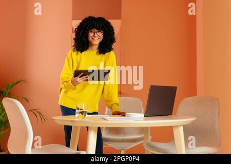 Giovane donna d'affari sorridente in piedi con un tablet PC davanti alla parete arancione Foto Stock