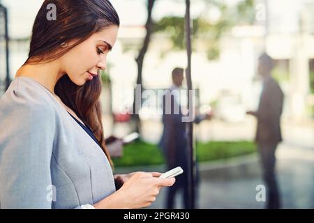 Rimanere connessi mentre ci si aspetta di entrare. una bella giovane donna d'affari che usa un cellulare fuori. Foto Stock