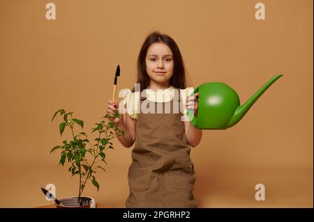 Adorabile bambina sorridente in possesso di annaffiatura lattina e pala da giardino, impara il giardinaggio, isolato su sfondo beige Foto Stock