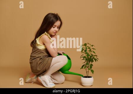 Bella bambina a tutta lunghezza in abito cachi, versando un po' d'acqua in un vaso di fiori con pianta di peperoncino in fiore Foto Stock