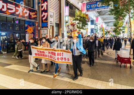 I manifestanti giapponesi marciano attraverso una galleria di negozi nella città di Kumamoto la sera con un banner sul davanti e diversi manifestanti in possesso di cartelli. La manifestazione è contraria all'aumento della spesa per la forza di difesa civile. Foto Stock