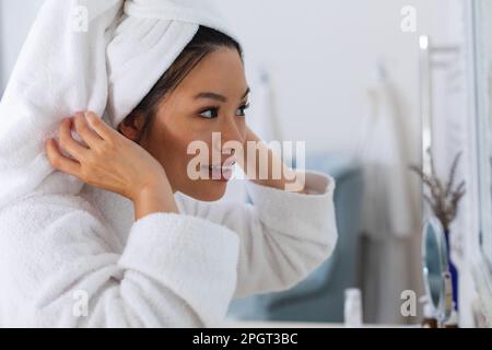 Felice donna asiatica che indossa accappatoio e asciugamano sulla testa guardando se stessa nello specchio del bagno. Stile di vita, cura di sé, salute e bellezza. Foto Stock
