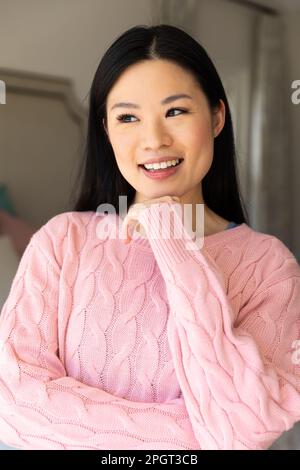 Donna asiatica felice che indossa un maglione rosa sorridente e che guarda lontano in camera da letto a casa. Stile di vita, tempo libero e relax. Foto Stock